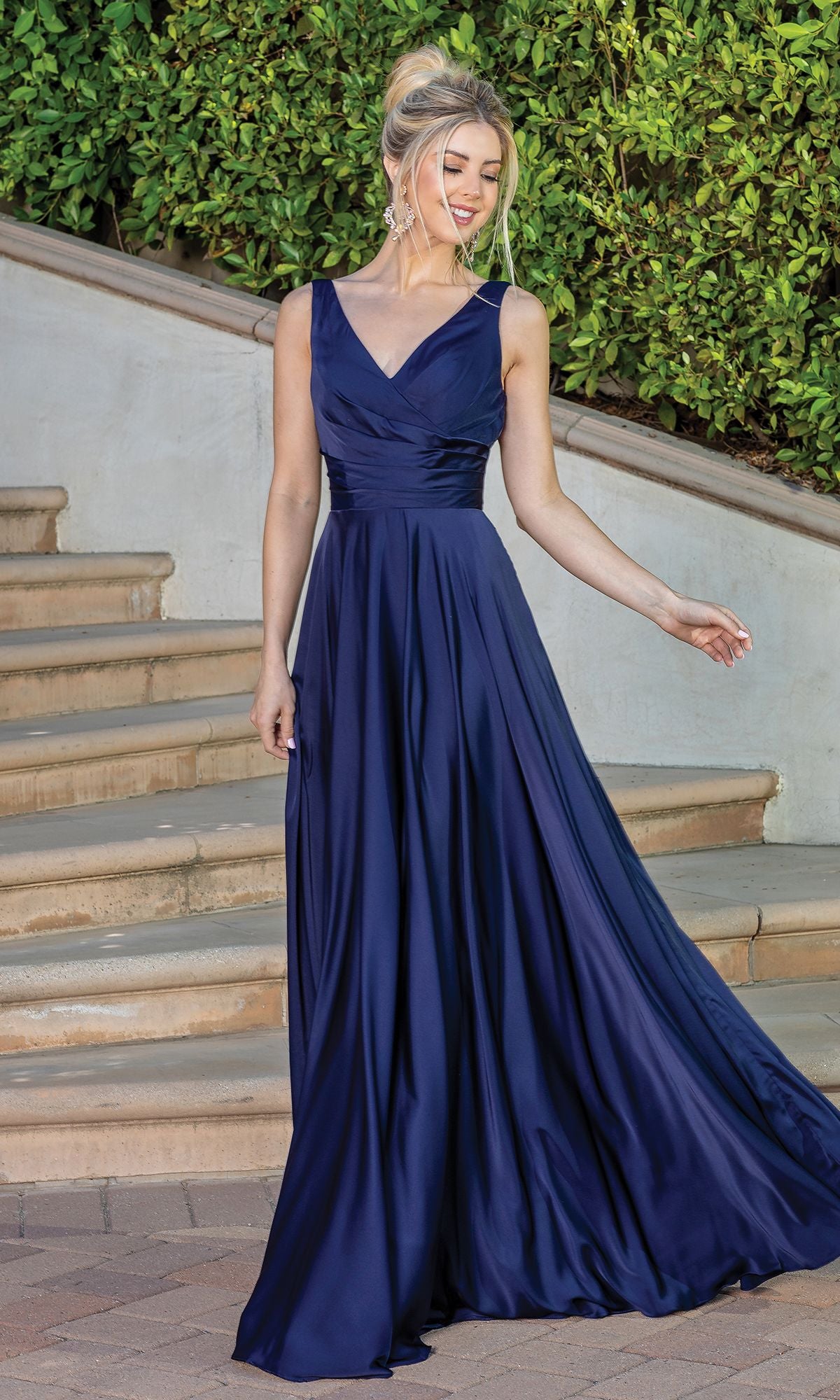 A woman in a navy blue Wrap-Bodice Long A-Line Prom Dress with Corset stands outdoors by stone steps and greenery, gazing to the side while holding part of her sleeveless, floor-length gown.